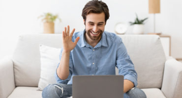 Video Call. Man Waving Hand To Laptop Screen Communicating Online Sitting On Sofa At Home. Guy Gesturing Hello To Computer Having Video Conference. Modern Distant Communication Concept